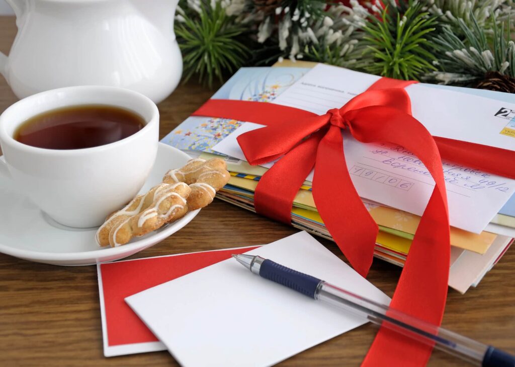 Stack of holiday cards tied with a red ribbon beside a cup of tea, cookies, a pen and a blank card.