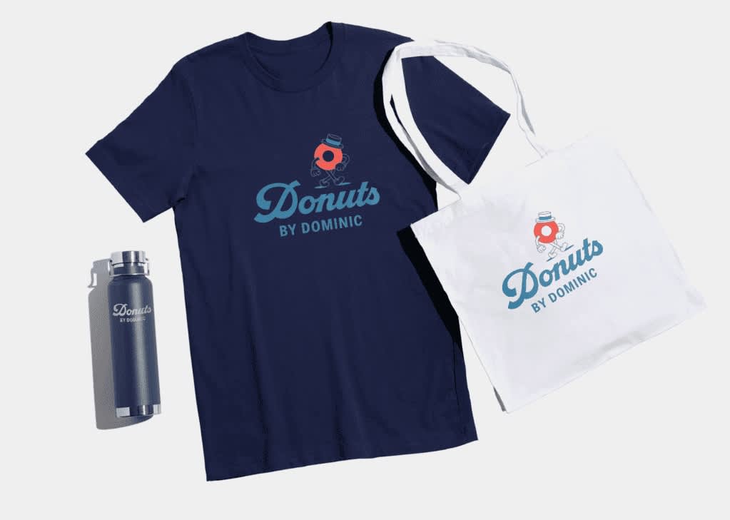 Selection of &ldquo;Donuts by Dominic&rdquo; logo merchandise, including a dark blue T-shirt, a white tote bag, and a navy thermal tumbler. Merch features light blue script text and a dapper red donut character.