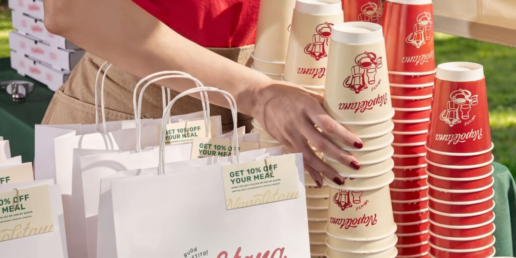 A hand holding a stack of branded paper cups next to a paper takeout bag.