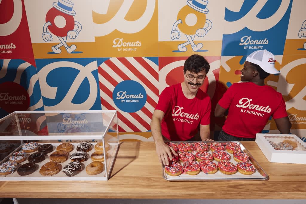 Two employees behind the counter of a donut shop wearing matching red T-shirts with white font &ldquo;Donuts BY DOMINIC.&rdquo;