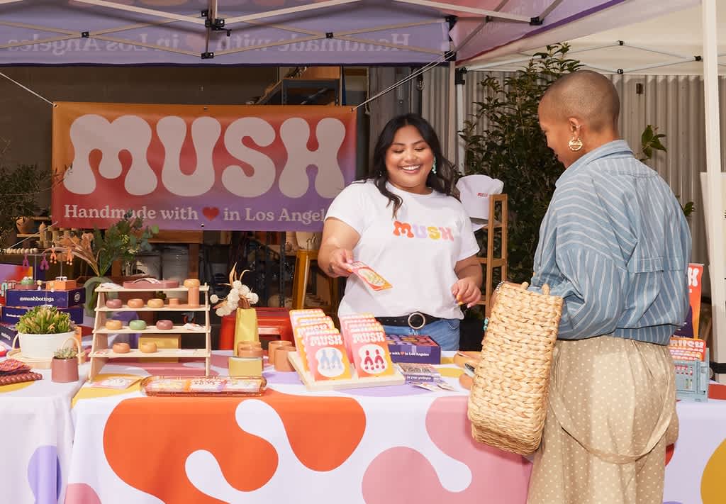 Person wearing a branded white T-shirt while serving a customer at a colorful outdoor market stall.