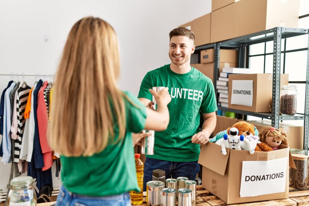 Two volunteers wearing green shirts while sorting donations, with boxes, food and clothing in the room.