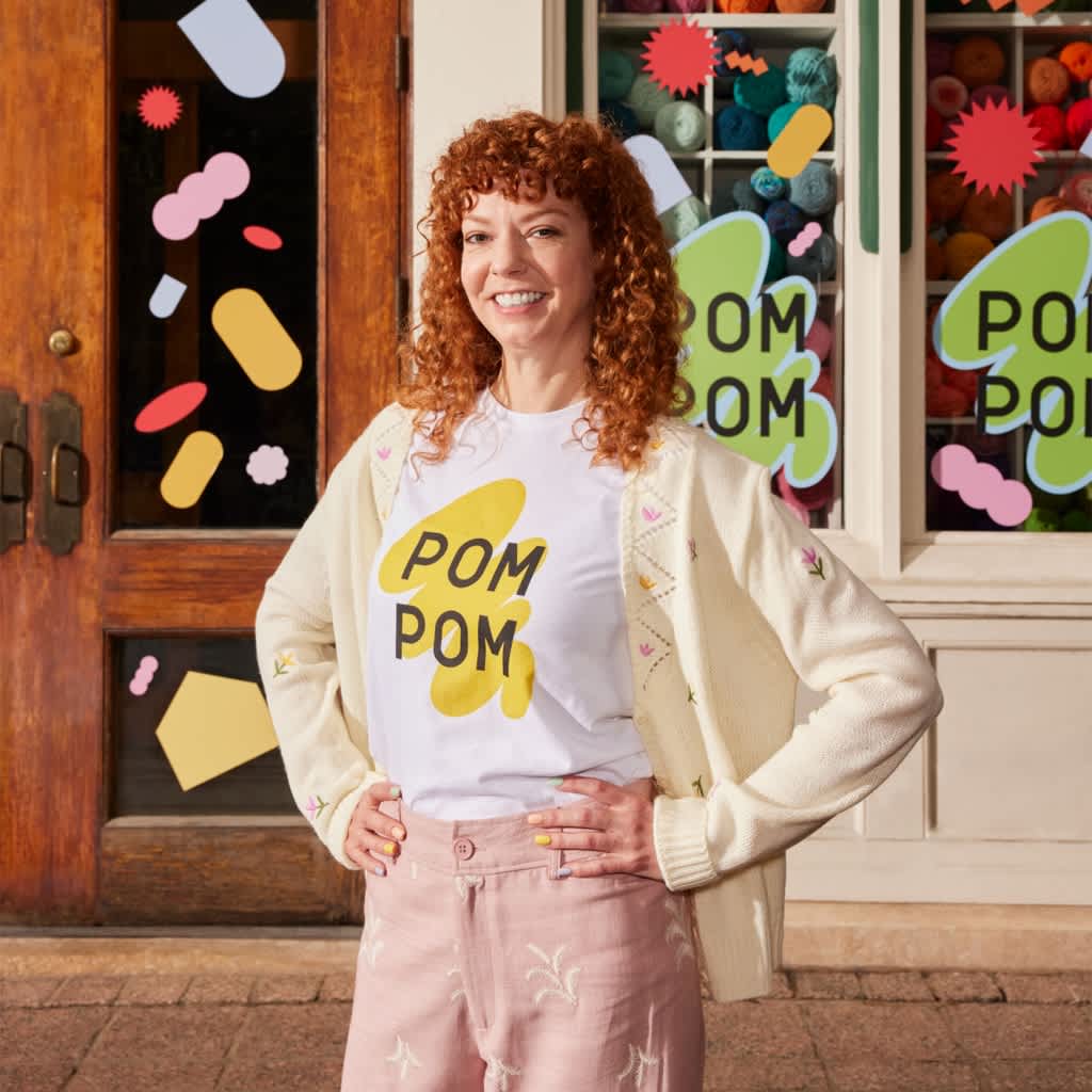 Small business owner in front of craft store wearing white T-shirt with large yellow paint wipe design with font &ldquo;POM POM.&rdquo;
