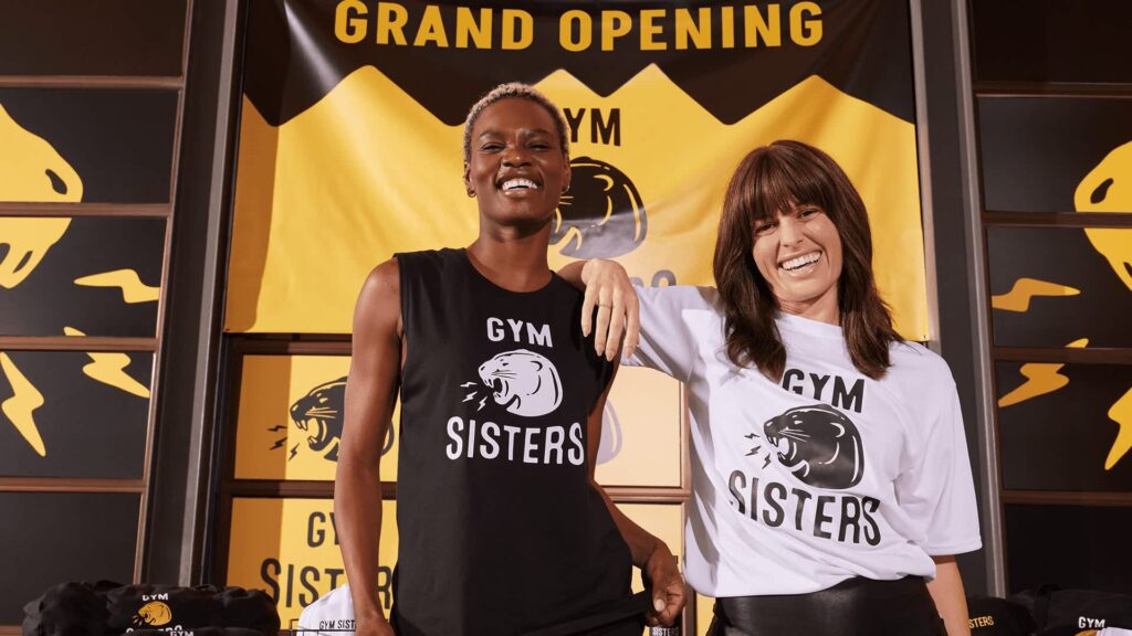 Two women in branded T-shirts stand in front of the brand&rsquo;s grand opening store front. The brand reads &ldquo;gym sisters&rdquo; with a roaring lioness logo.