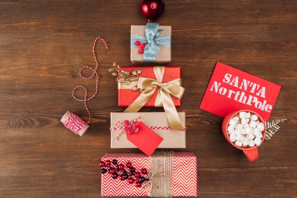 Wrapped gift boxes arranged in a Christmas tree shape on a wooden surface, with bows, tags and holiday decorations.