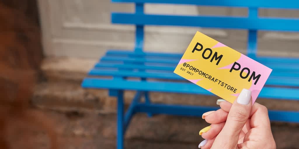 Hand holding up bright pink and yellow business card with chunky black “Pom Pom - @pompomcraftstore” text.