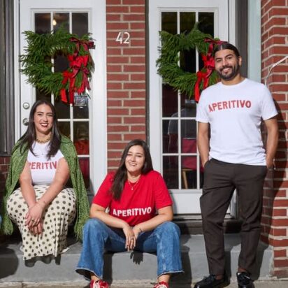 Four people wearing matching branded T-shirts while sitting and standing outside a brick storefront.