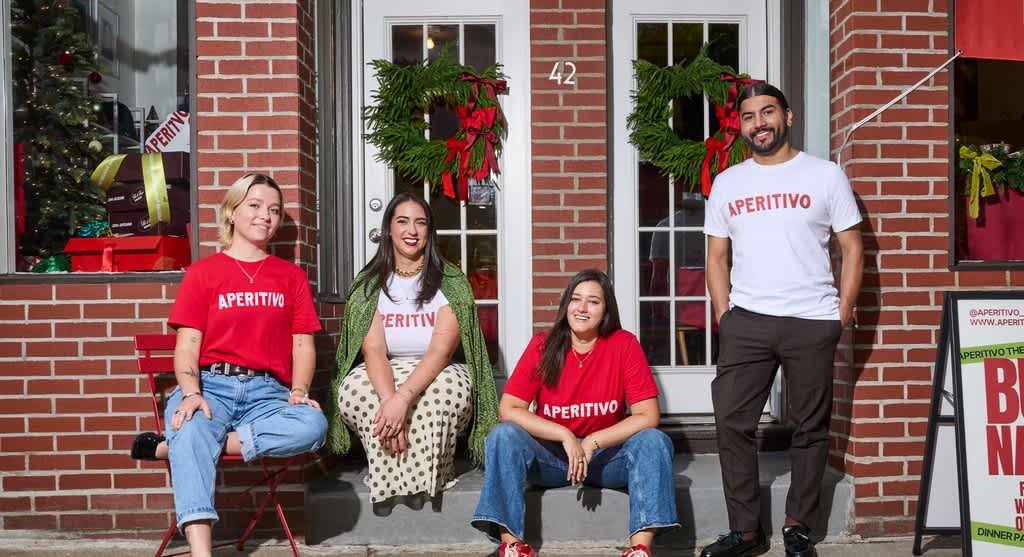 Four people wearing matching branded T-shirts while sitting and standing outside a brick storefront.