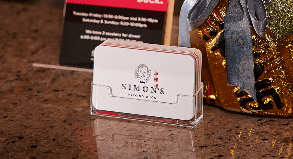 Stack of white rounded edge business cards with red Chinese characters, black logo of face and black “Simon’s Peiking Duck” text, in a clear acrylic card holder.