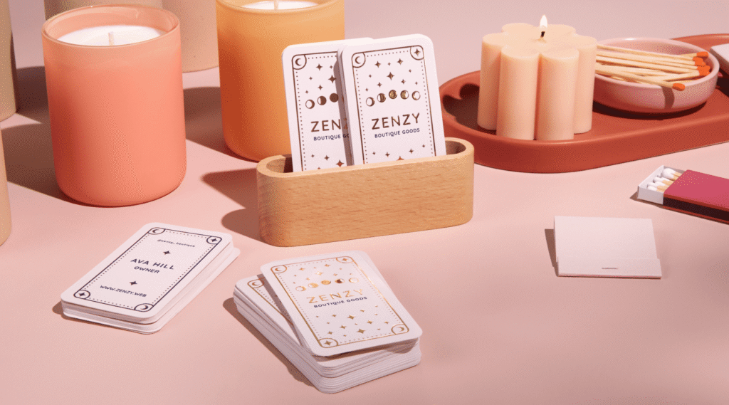 Stack of rounded edge white business cards with gold raised foil design and “Zenzy Boutique Goods” text in a wooden card holder next to selection of candles.