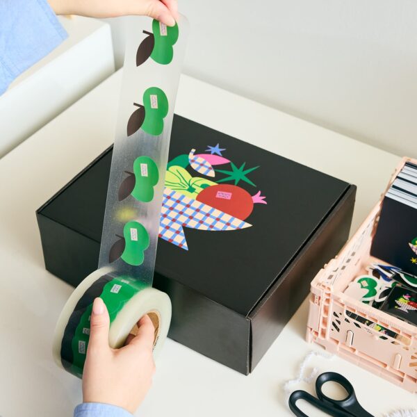 Person unrolling custom packaging tape with green fruit illustrations beside a black mailer box decorated with colorful branded graphics.