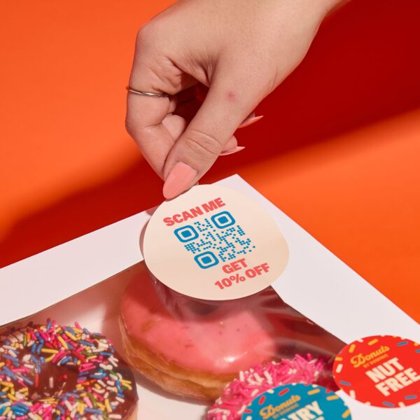 Person applying a round promotional QR code sticker offering 10% off to a donut box filled with colorful frosted donuts.