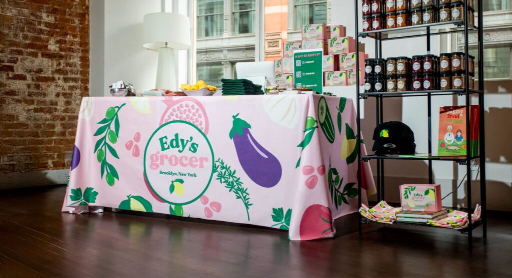 A small business grocery stand with table banner and shelves of products, merch and branded product boxes.