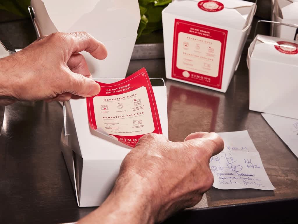 Small business owner attaching red and white custom labels onto small white takeout boxes.