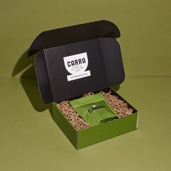Open green and black CORRA branded mailer box containing a green product pouch nestled in brown crinkle paper, with a printed logo inside the lid.