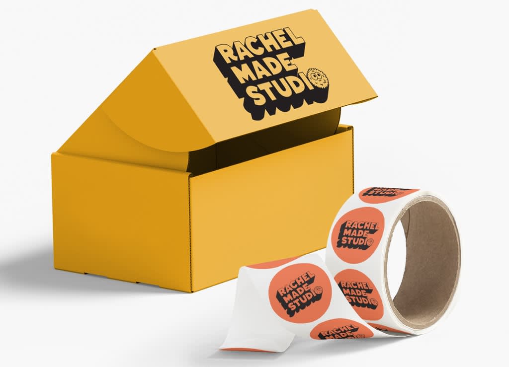 Yellow branded cardboard package with black “Rachel Made Studio” text and a roll of circular custom stickers with the same branded text.