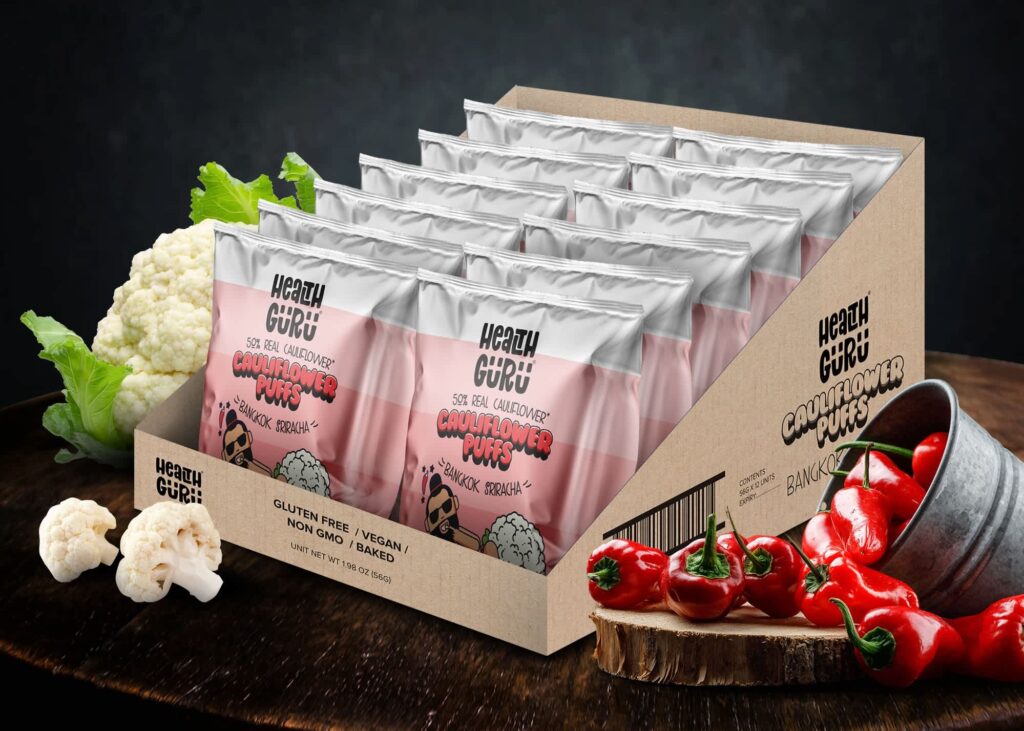 Pink Health Guru cauliflower puff snack bags displayed in a cardboard tray beside cauliflower and red peppers.