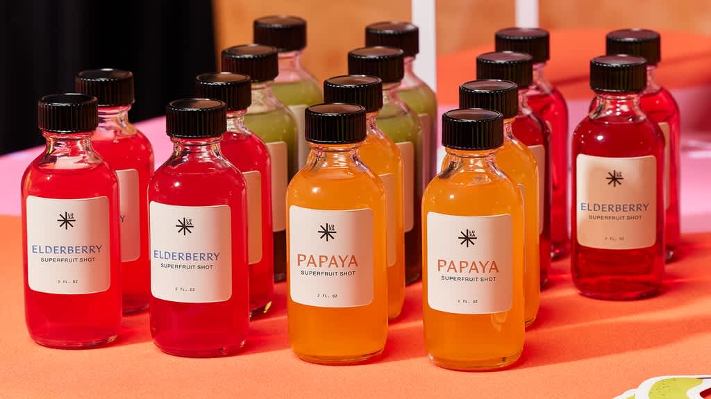 Rows of colorful superfruit shot bottles labeled Elderberry and Papaya on an orange display surface.