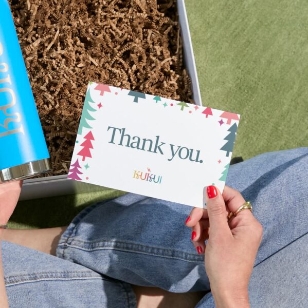 Person holding a KUKUI branded thank-you postcard with a colorful Christmas tree design beside a gift box filled with brown crinkle paper.