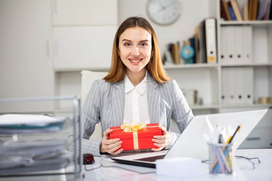 Elegant woman in an office holding a red gift box with a golden ribbon, smiling – the perfect employee appreciation gift.