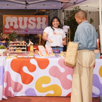 Outdoor fair stand showcasing signage in retail stores, with coordinated banner, vertical flag, A-sign and smiling vendor and customer.