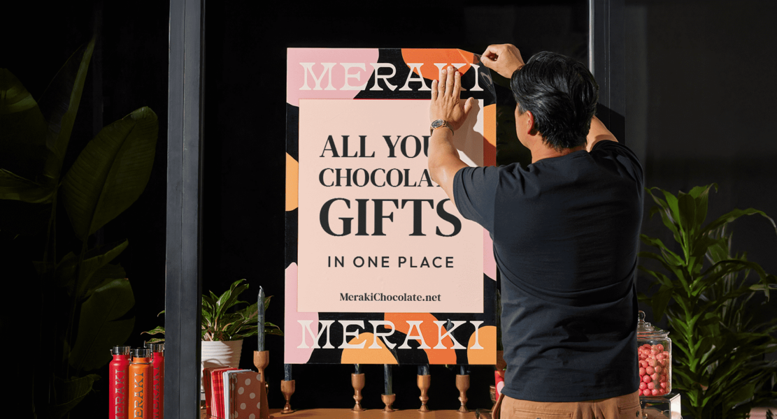 Store owner applying adhesive poster to storefront window surrounded by branded products and potted plants — signage for business in action.