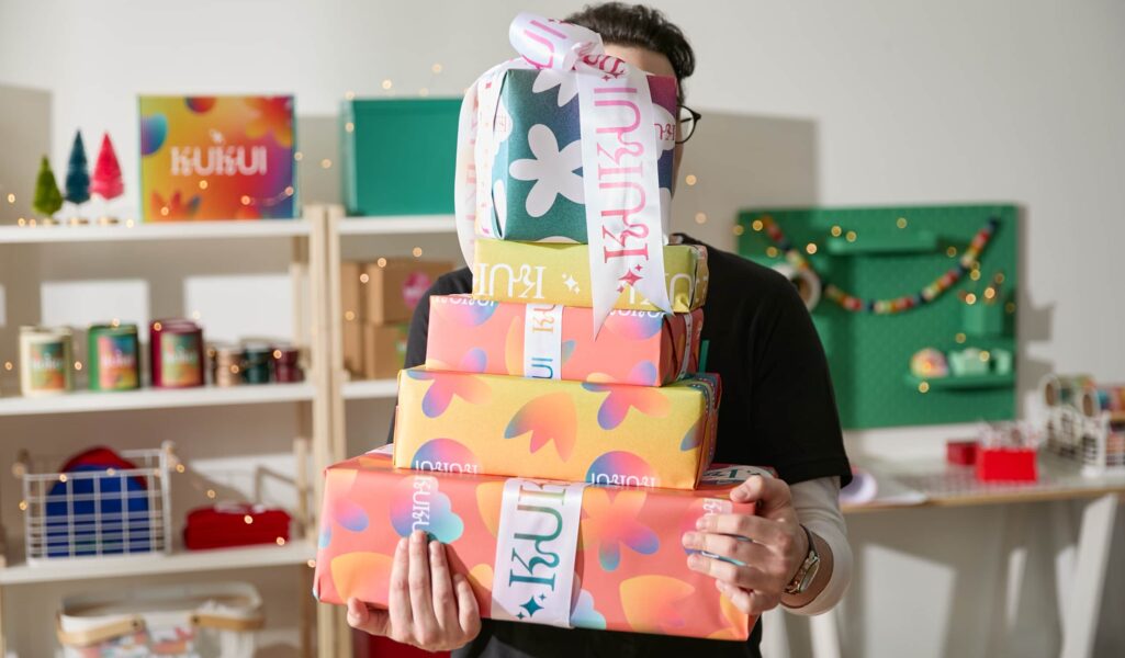 Man holding a stack of branded corporate Christmas gifts with festive lights and matching merchandise in the background