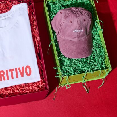 Three products in festive packaging, with the visible branding "Apertivo", are laid out side by side on a red table: A beanie, a thsirt and a cap