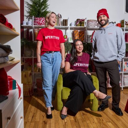 Business holiday prep example: three smiling people wearing branded Aperitivo accessories, t-shirts, and promotional materials in the background.