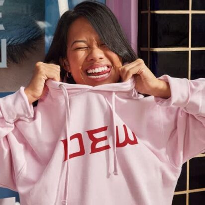 Woman smiles as she pulls a pink branded hoodie down over her head