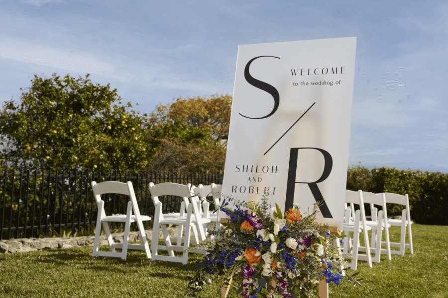 Wedding welcome sign with colorful floral arrangement on a wooden easel, set on a lawn with white ceremony chairs.