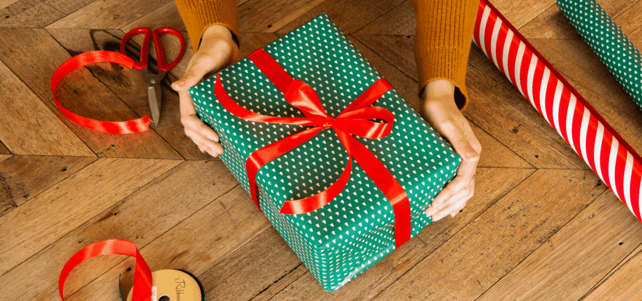 Zwei Hände halten ein grün verpacktes Geschenk mit roter Schleife auf einer Holzoberfläche.