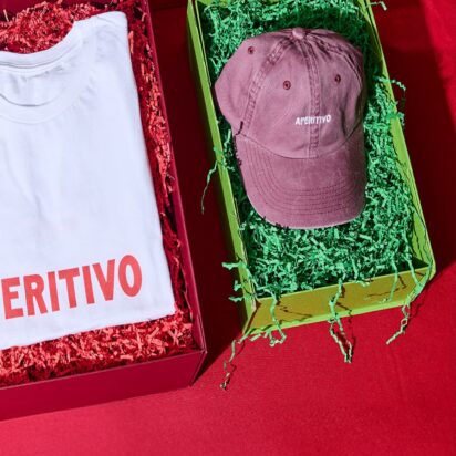 Three products in festive packaging, with the visible branding "Apertivo", are laid out side by side on a red table: A beanie, a thsirt and a cap