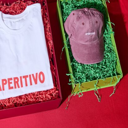 Three products in festive packaging, with the visible branding "Apertivo", are laid out side by side on a red table: A beanie, a thsirt and a cap
