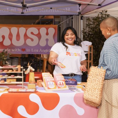 A small business owner handing a shopping bag to a customer at a MUSH-branded outdoor market booth with handmade products.