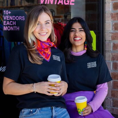 Two women wearing branded T-shirts and holding takeout drink cups in front of their store window.
