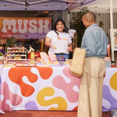 Outdoor fair stand showcasing signage in retail stores, with coordinated banner, vertical flag, A-sign and smiling vendor and customer.