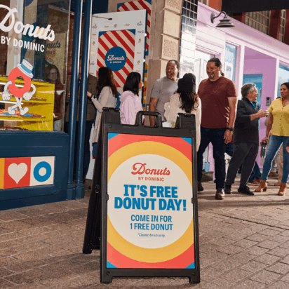 Colorful A-frame sign outside a donut shop with people lined up to enter.